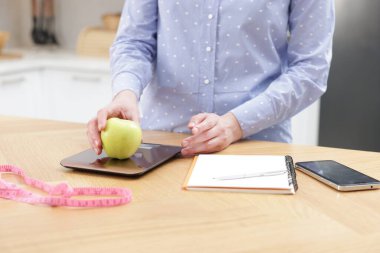 Mutfak terazisinde elmayı tartan kadın, kapalı masa, yakın plan.