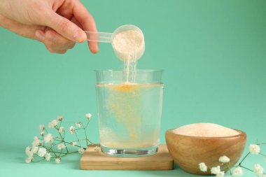 Kadın bardağa kolajen tozu ekliyor. Turkuaz arka planda su var. Yakın plan.