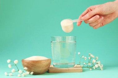 Kadın bardağa kolajen tozu ekliyor. Turkuaz arka planda su var. Yakın plan.