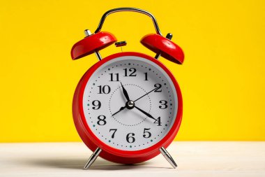 Sarı arka planda beyaz ahşap masada kırmızı bir çalar saat.