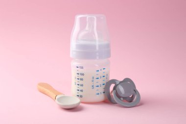 Pembe arka planda süt, kaşık ve emzik ile beslenme şişesi