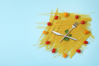 Çiğ spagetti, kiraz domatesleri ve açık mavi arka planda çatal bıçakla yapılmış saat. Metin için boşluk