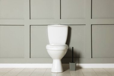 Ceramic toilet bowl and brush near grey wall indoors