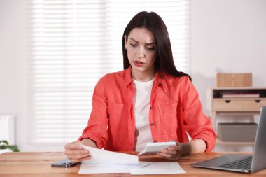 Bilgisayarı, akıllı telefonu ve hesap makinesi olan bir kadın kapalı bir masada faturaları ödüyor.