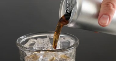 Kadın teneke kutudan bardağa soda dolduruyor. Gri arka planda buz küpleri var. Yakın plan.