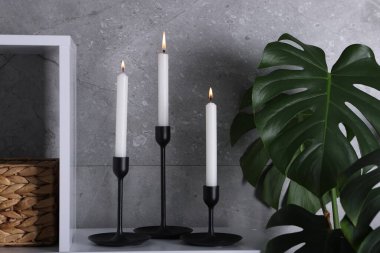 Rafta yanan mumlarla süslü şamdanlar ve kapalı alanda gri duvarın yanında bitki yetiştirme.