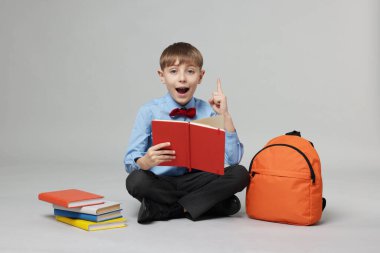 Sırt çantası açık gri arka planda kitap okuyan sevimli bir okul çocuğu.