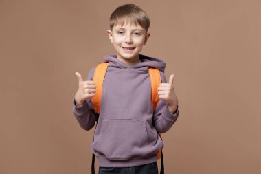 Sırt çantalı yakışıklı okul çocuğu kahverengi arka planda baş parmak gösteriyor.
