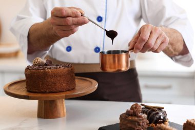 Bir kepçe erimiş çikolatayla yaşlı pasta şefi mutfaktaki masada pasta yapıyor, yakın plan.