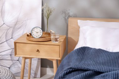 Komodinin yanında çalar saat, vazoda çiçekler ve yatağın yanında bir bardak su.