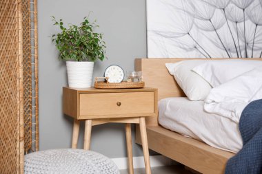 Yatağın yanında ev bitkisi, çalar saat ve yatağın yanında bir bardak su olan komodin.