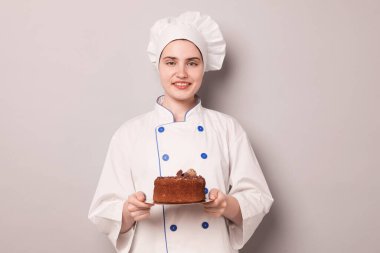 Üniformalı pastacı açık gri arka planda lezzetli çikolatalı pasta tutuyor.
