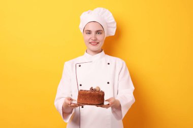 Üniformalı şekerlemeci turuncu arka planda lezzetli çikolatalı pasta tutuyor.