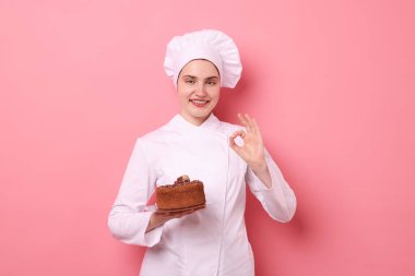 Üniformalı bir şekerci lezzetli çikolatalı pasta tutuyor ve pembe arka planda güzel bir jest yapıyor.