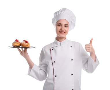 Üniformalı mutlu pastacı elinde çilekli lezzetli profiteroller tutuyor ve beyaz arka planda baş parmak gösteriyor.