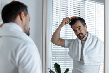 Yakışıklı bir adam saçını aynanın yanında tarıyor.