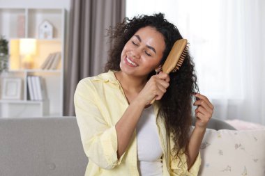 Gülümseyen genç bir kadın evde kıvırcık saçlarını tarıyor.