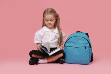 Sırt çantalı liseli kız pembe arka planda kitap okuyor.