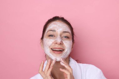 Spa günü. Güzel bir kadın pembe arka planda yüzüne maske takıyor.