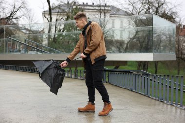 Kırık şemsiyeli bir adam açık havada yağmur altında rüzgarla savaşıyor.