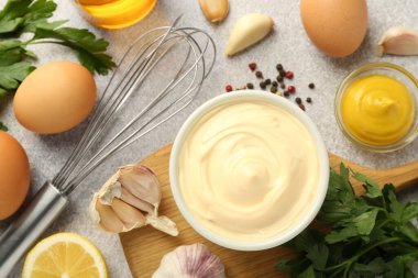 Lezzetli mayonez sosu kasede, çırpı ve malzemeler gri masada, düz yerde.