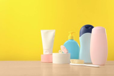 Bebek kozmetik ürünleri ve banyo aksesuarları ahşap masada, sarı arka planda. Metin için boşluk