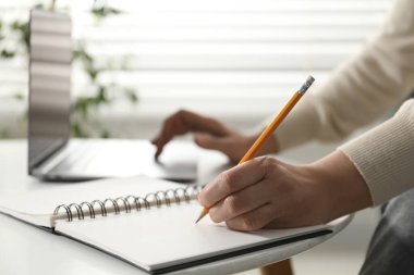 İnternette bilgisayar kullanarak öğrenen ve içerideki beyaz masada notlar alan bir kadın. Kendi kendine çalışma.