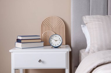 Yatağın yanında kitap, dekor ve alarmlı bir masa var.