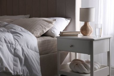 Yatağın yanında kitap, lamba ve bir bardak su olan komodin.