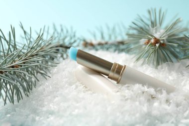 Nemlendirici dudak kremi ve yapay kar üzerindeki köknar ağacı dalları açık mavi arka planda, yakın plan. Kış bakımı