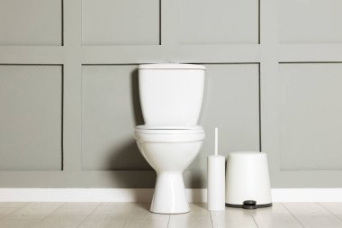 Seramik klozet, fırça ve çöp tenekesi kapalı alanda gri duvarın yanında.