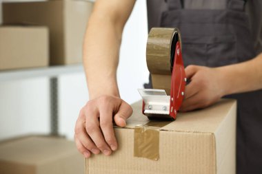 Karton kutuyu kapalı bir yere kapatmak için yapıştırıcılı bant kullanan bir adam.