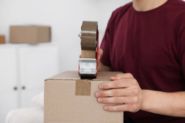 Karton kutuyu kapalı bir yere kapatmak için yapıştırıcılı bant kullanan bir adam.