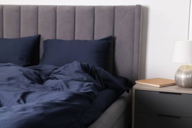 Evinde koyu mavi ipek çarşaflar ve komodinli bir yatak.