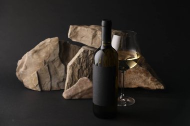 Bir şişe şarap, şarap kadehi ve siyah arka planda kayalar.