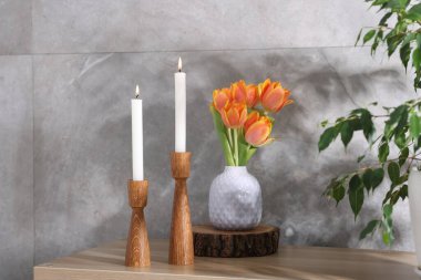 Havalı şamdanlar, yanan mumlar, güzel çiçekler ve kapalı alandaki gri mermer duvarın yanındaki ahşap masa üzerindeki ev bitkisi.