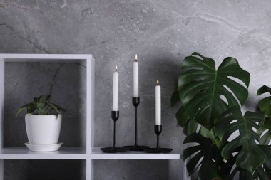 Rafta yanan mumlarla süslü şamdanlar ve kapalı alanda gri duvarın yanında bitkiler.