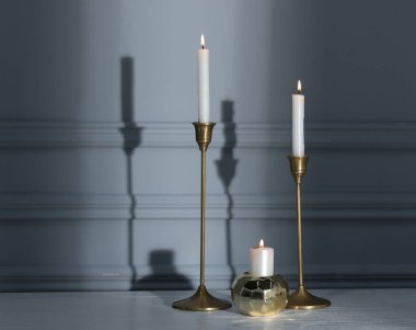 Gri duvarın yanındaki beyaz masada yanan mumlarla süslü şamdanlar.