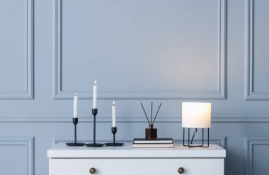 İçinde yanan mumlar, kitaplar ve diğer dekorlar olan şık şamdanlar kapalı alandaki gri duvarın yanındaki beyaz çekmecelerin üzerine.