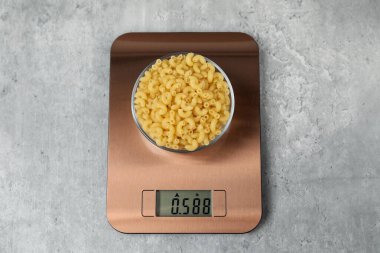 Modern electronic kitchen scale with bowl of raw pasta on grey table, top view