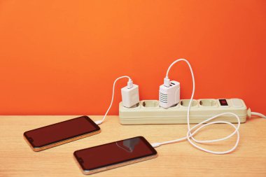 Kablolu USB adaptörleri elektrik kablosuna takılı turuncu duvarın yanındaki ahşap masada akıllı telefonları şarj ediyor.
