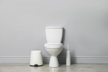 Seramik klozet, fırça ve çöp tenekesi kapalı alanda gri duvarın yanında.