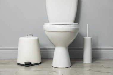 Seramik klozet, fırça ve çöp tenekesi kapalı alanda gri duvarın yanında.