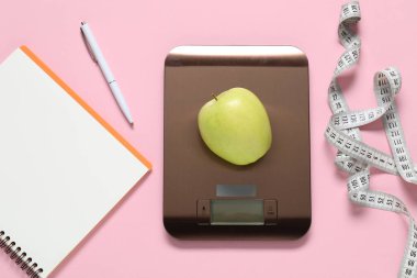 Elektronik mutfak ölçeği, elma, mezura, pembe arka planda not defteri ve kalem, düz konum. Kalori kontrolü