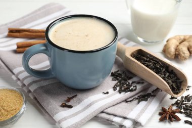 Aromatik Masala çayı fincanda, baharatlar, kuru yapraklar, beyaz ahşap masada süt ve kahverengi şeker, yakın plan.