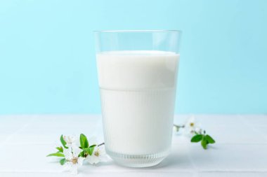 Açık mavi arka planda beyaz fayanslı masada bir bardak taze süt ve çiçek.