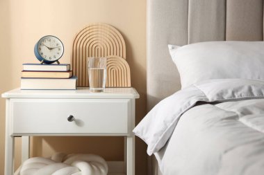 Yatağın yanında kitap, dekor, bir bardak su ve çalar saat olan komodin.
