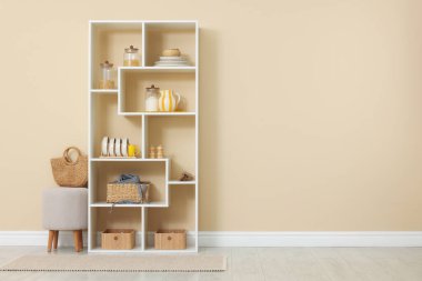 Shelving unit with dishware, jars of different products and other stuff near beige wall indoors, space for text
