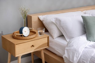 Komodinin yanında çalar saat, vazoda çiçekler ve yatağın yanında bir bardak su.