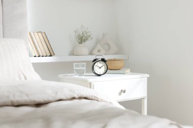 Alarm saati, bir bardak su, hasır kutu ve yatağın yanında kitap olan komodin.